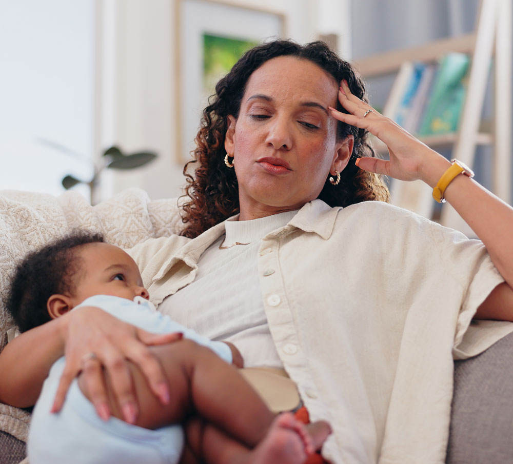 Tired woman holds baby, sitting on couch.