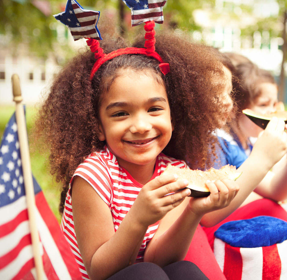 Child smiling, eating watermelon, wearing patriotic headband