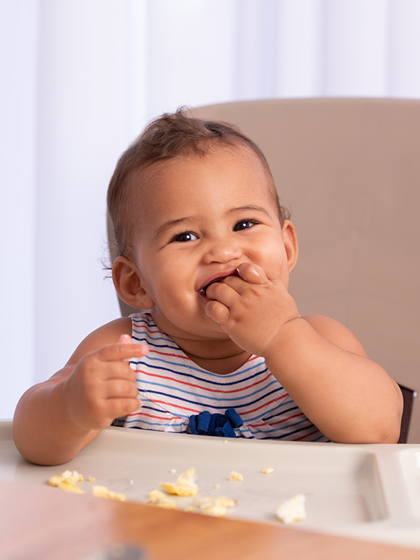 Smiling baby eating scrambled eggs in high chair.