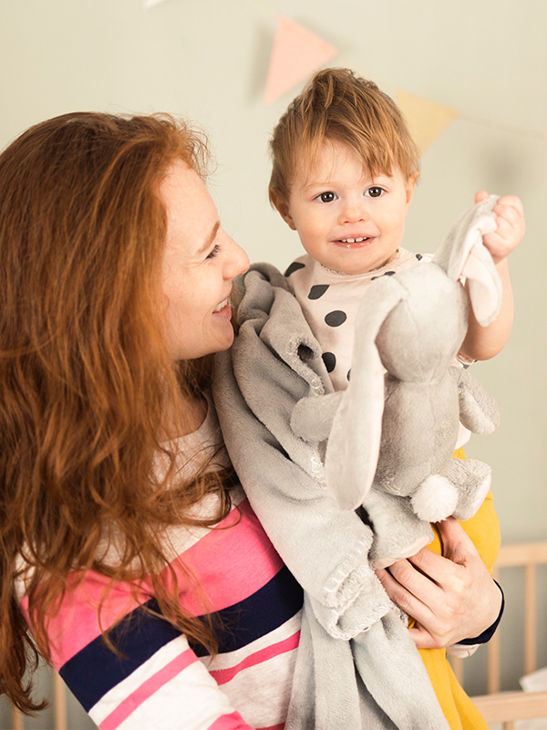 Woman holding smiling toddler with gray plush toy.