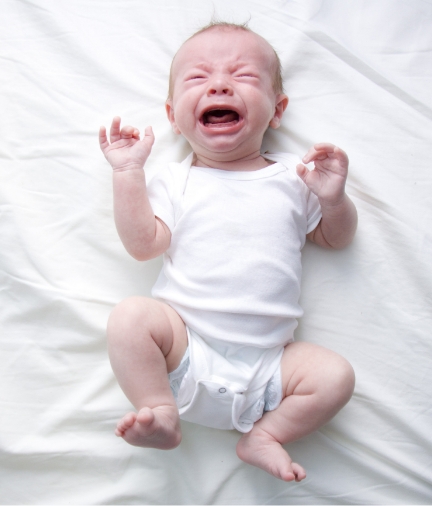 Crying baby lying on white bed sheet.
